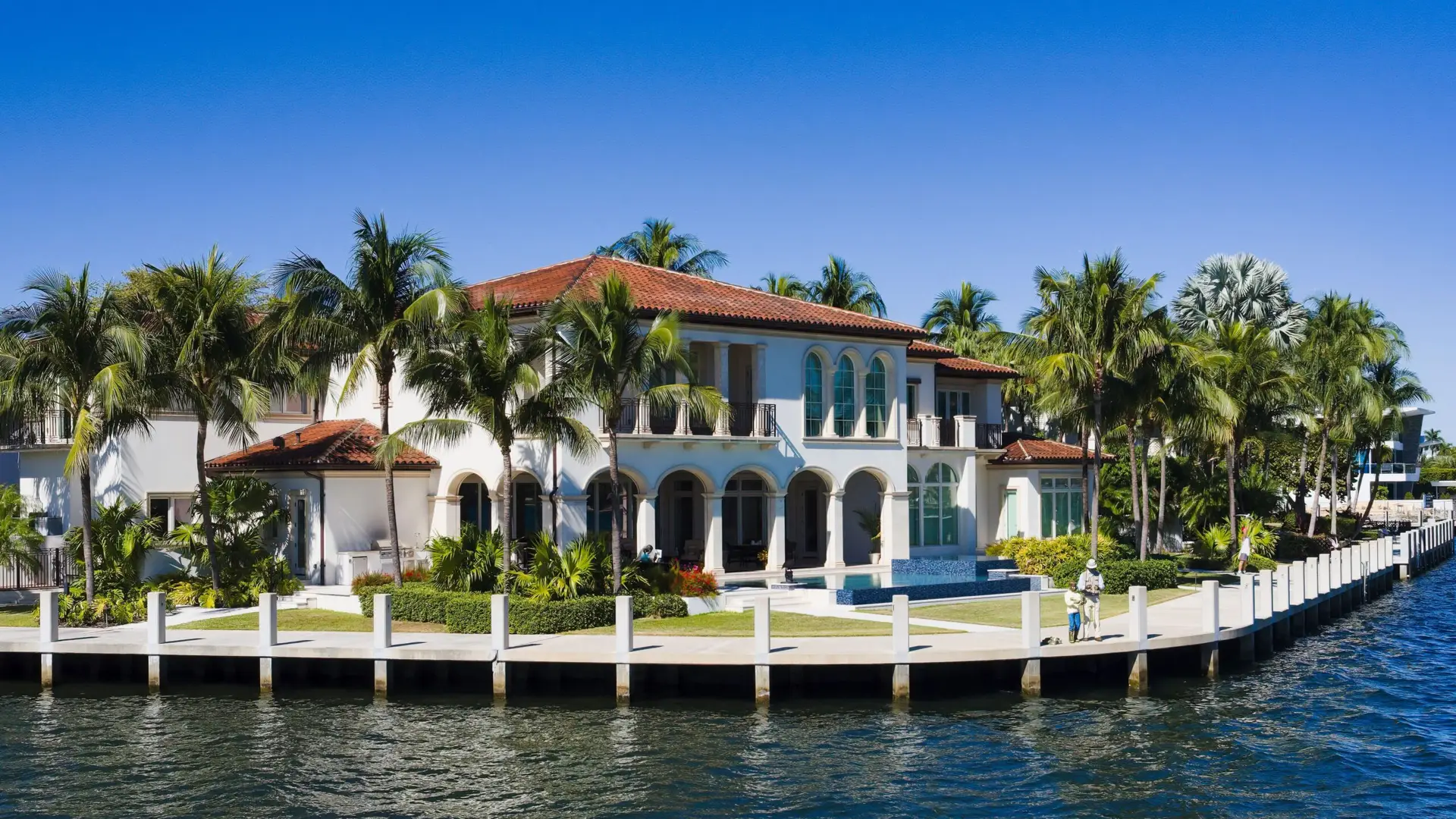 Canal-front estate with concrete seawall cap, dock pilings, pool, and palms on a South Florida waterway