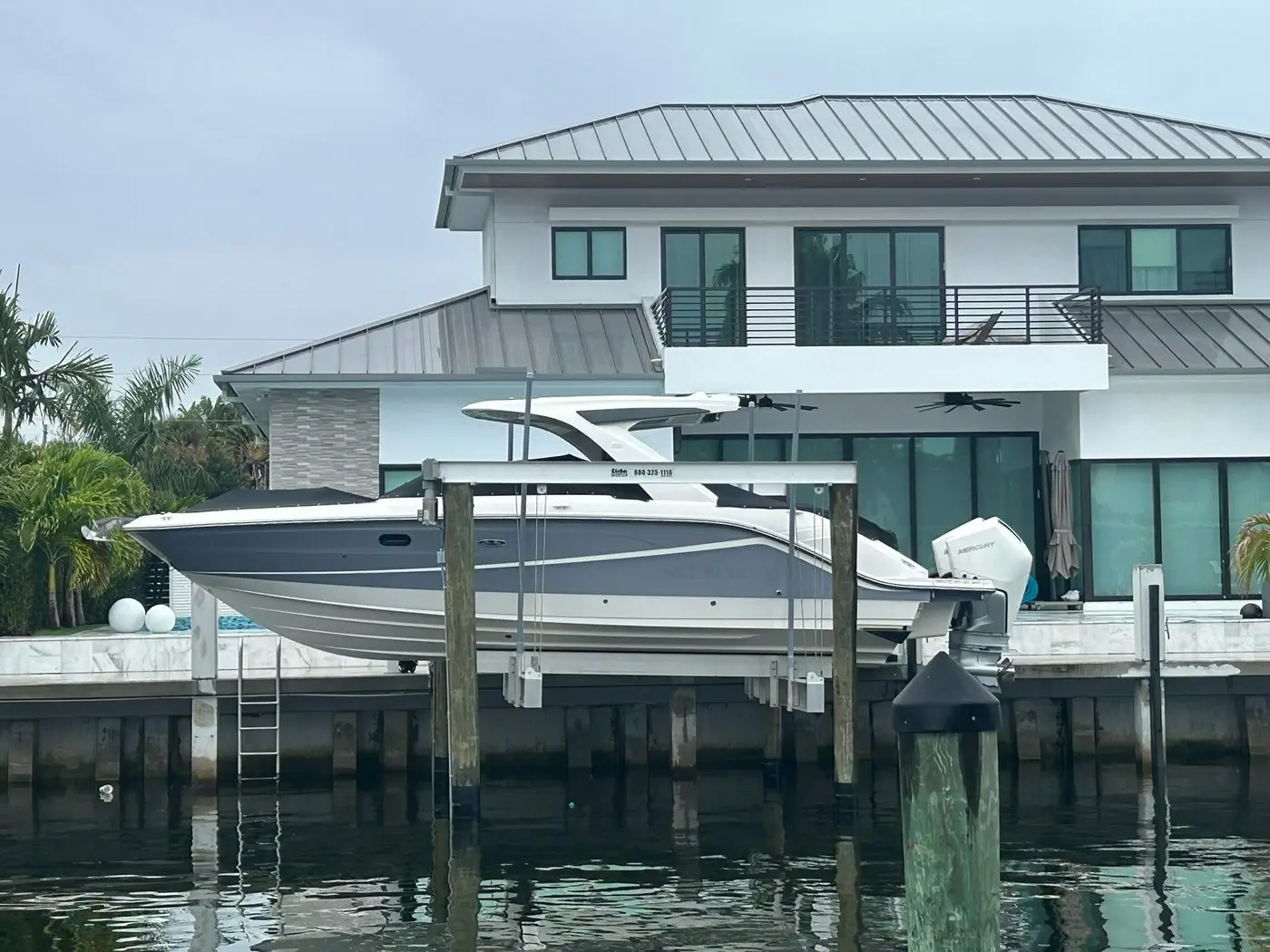 Boat raised on a lift beside a private dock, with pilings and seawall in front of a modern waterfront home